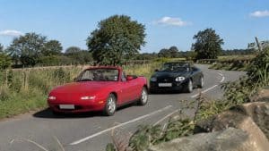 Mazda MX-5 NA rouge suivie d'un MGF noir sur une route de campagne ensoleillée