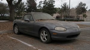 Mazda MX-5 NB grise à l'abandon sur un parking, couverte de feuilles mortes, phares ternis