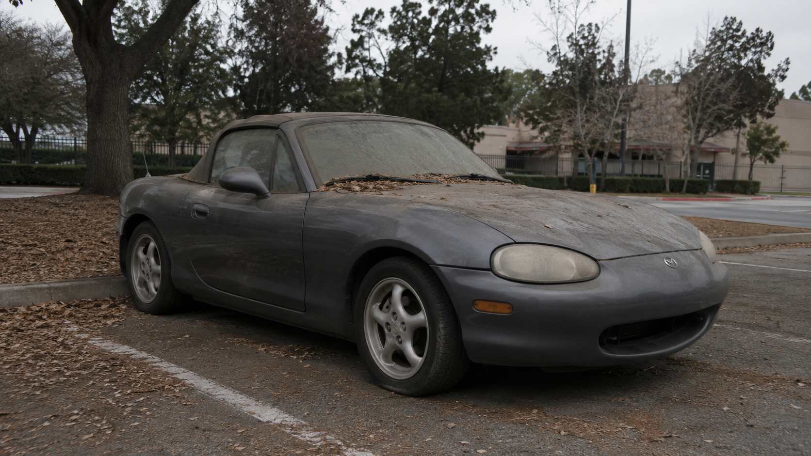 Mazda MX-5 NB grise a l'abandon sur un parking, couverte de feuilles mortes et phares ternis