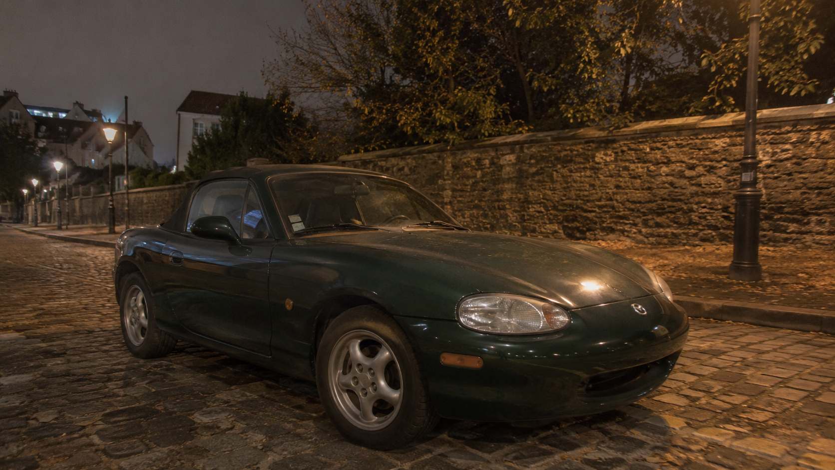 Mazda MX-5 NB verte garée de nuit dans une rue pavée éclairée par des lampadaires
