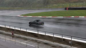 Photographie panoramique de 1672 x 941 px prise depuis les stands d'un circuit sous la pluie. Mazda MX-5 de quatrième génération ND, gris foncé métallisé, capote souple noire fermée, jantes peintes en noir, saisie de trois quarts arrière sur une piste entièrement détrempée : les roues arrière projettent une longue gerbe d'eau pulvérisée. Plaque d'immatriculation arrière de type britannique, à fond jaune. Le tracé décrit un virage à gauche bordé de vibreurs rouges et blancs, avec bandes de dégagement en herbe verte, glissières métalliques, grillage de sécurité, bloc de mousse rouge et noir, talus boisé en arrière-plan. Premier plan : muret de stand en béton surmonté d'une rambarde tubulaire, extincteur rouge accroché, panneau d'interdiction de fumer, voie des stands luisante marquée d'une ligne jaune et d'emplacements blancs. Ciel gris uniforme, lumière plate d'averse.