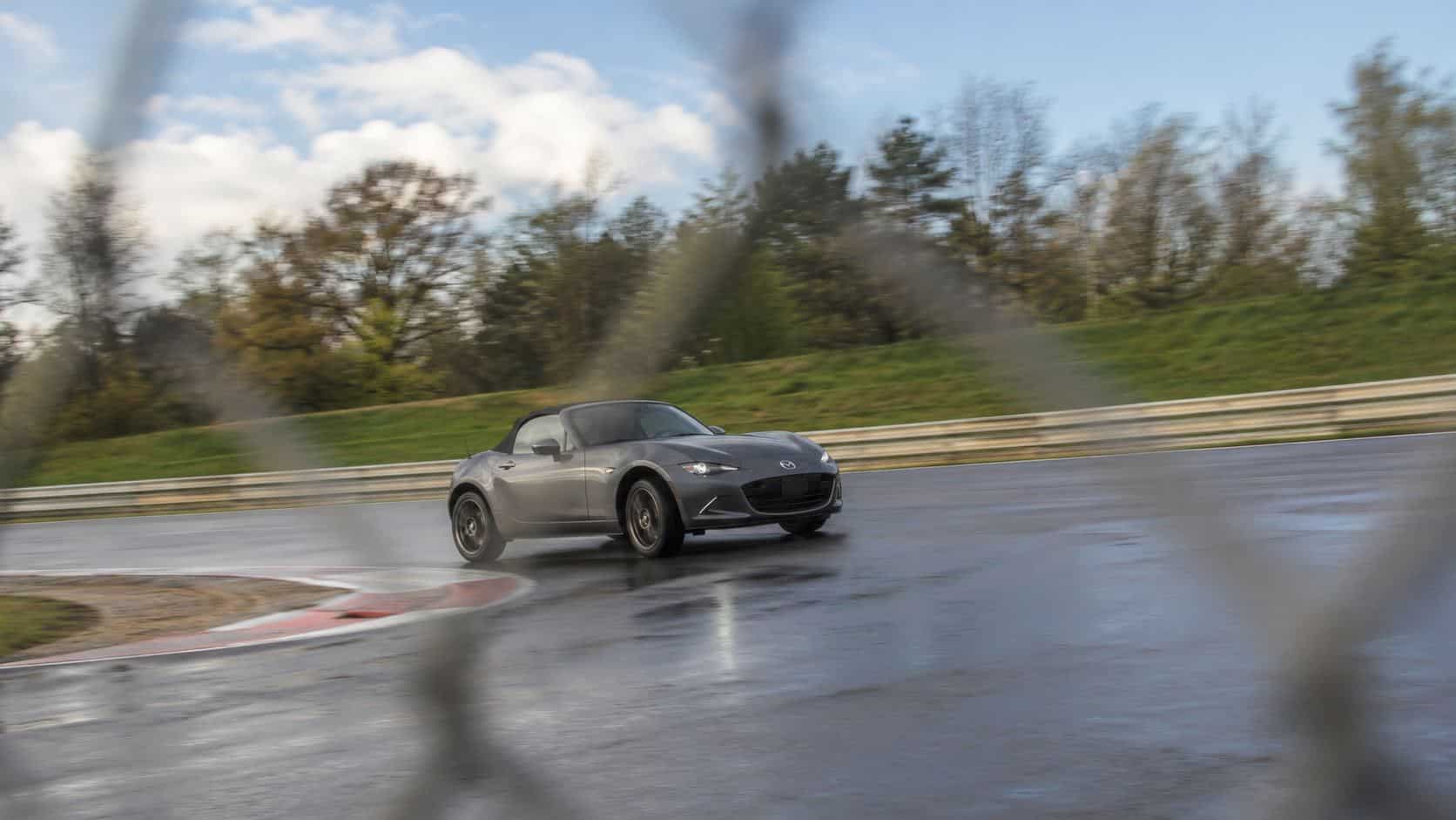 Mazda MX-5 ND grise capote fermée sur circuit mouillé, photographiée à travers un grillage flou