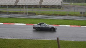 Mazda MX-5 ND grise capote fermée sur circuit détrempé sous la pluie, vibreurs rouge et blanc