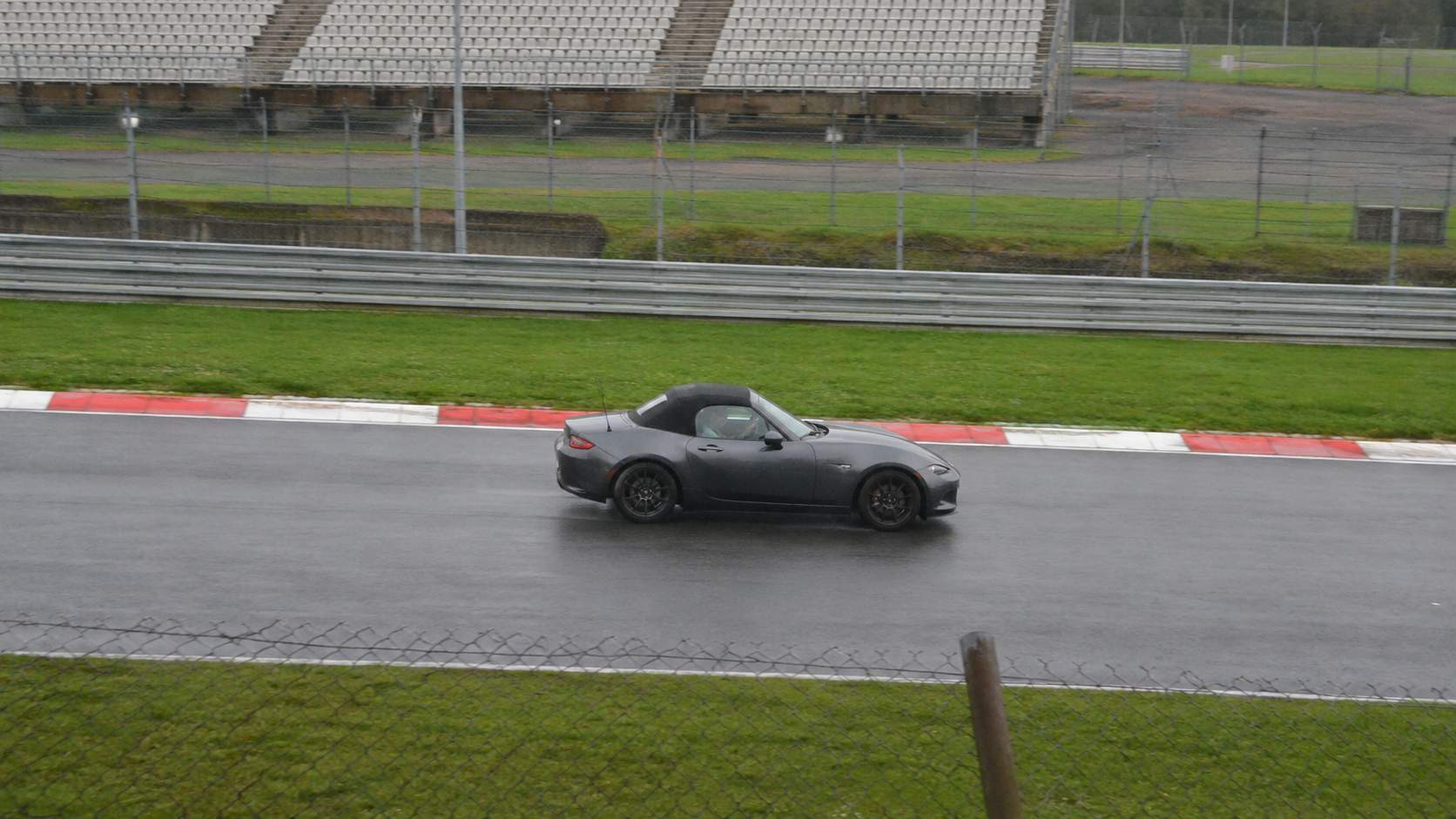 Mazda MX-5 ND grise capote fermée sur circuit détrempé sous la pluie, vibreurs rouge et blanc
