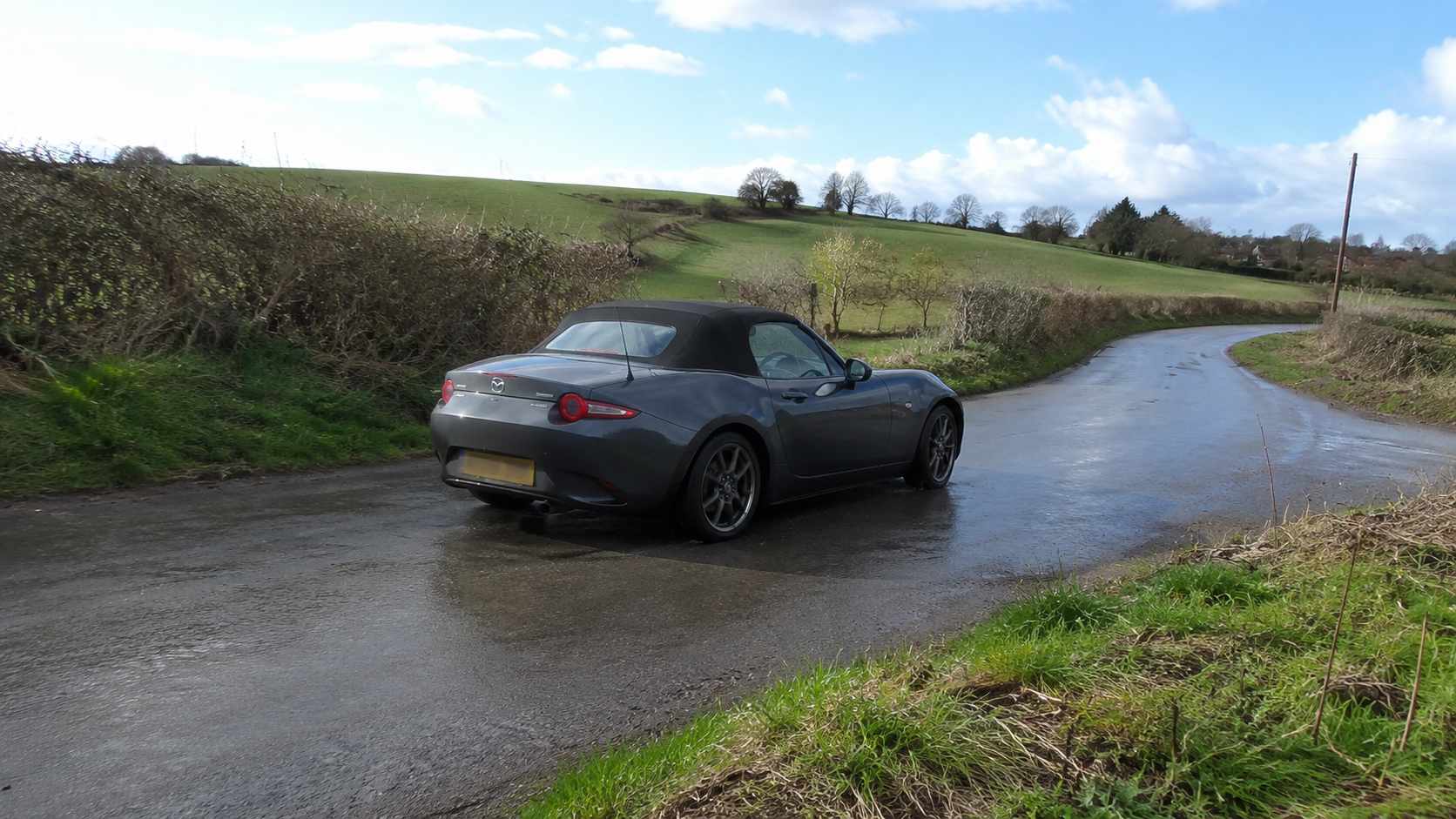 Mazda MX-5 ND gris anthracite sur une petite route de campagne mouillée bordée de haies
