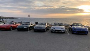 Photographie panoramique de 1672 x 941 px prise au coucher du soleil sur un parking bitumé en bord de mer. Cinq roadsters Mazda MX-5 issus de quatre générations sont alignés de face, tous capote ouverte, sur des emplacements marqués au sol en blanc. De gauche à droite : une NA rouge vif à phares escamotables fermés, jantes sept branches argent et sellerie beige ; une NB vert foncé métallisé à phares fixes et intérieur beige ; une NB de seconde phase noire à jantes cinq doubles branches ; une NC gris argent métallisé, optiques en amande et large calandre noire, sellerie beige ; une ND bleu métallisé à calandre trapézoïdale noire, jantes peintes en noir et intérieur noir. Un sixième véhicule apparaît partiellement coupé au bord gauche du cadre. Arrière-plan : océan calme, ciel chargé de nuages orangés et gris, soleil bas à droite, rochers, végétation sèche, panneau de réglementation sur mât, conteneur à déchets noir, promeneurs sur la pointe rocheuse.