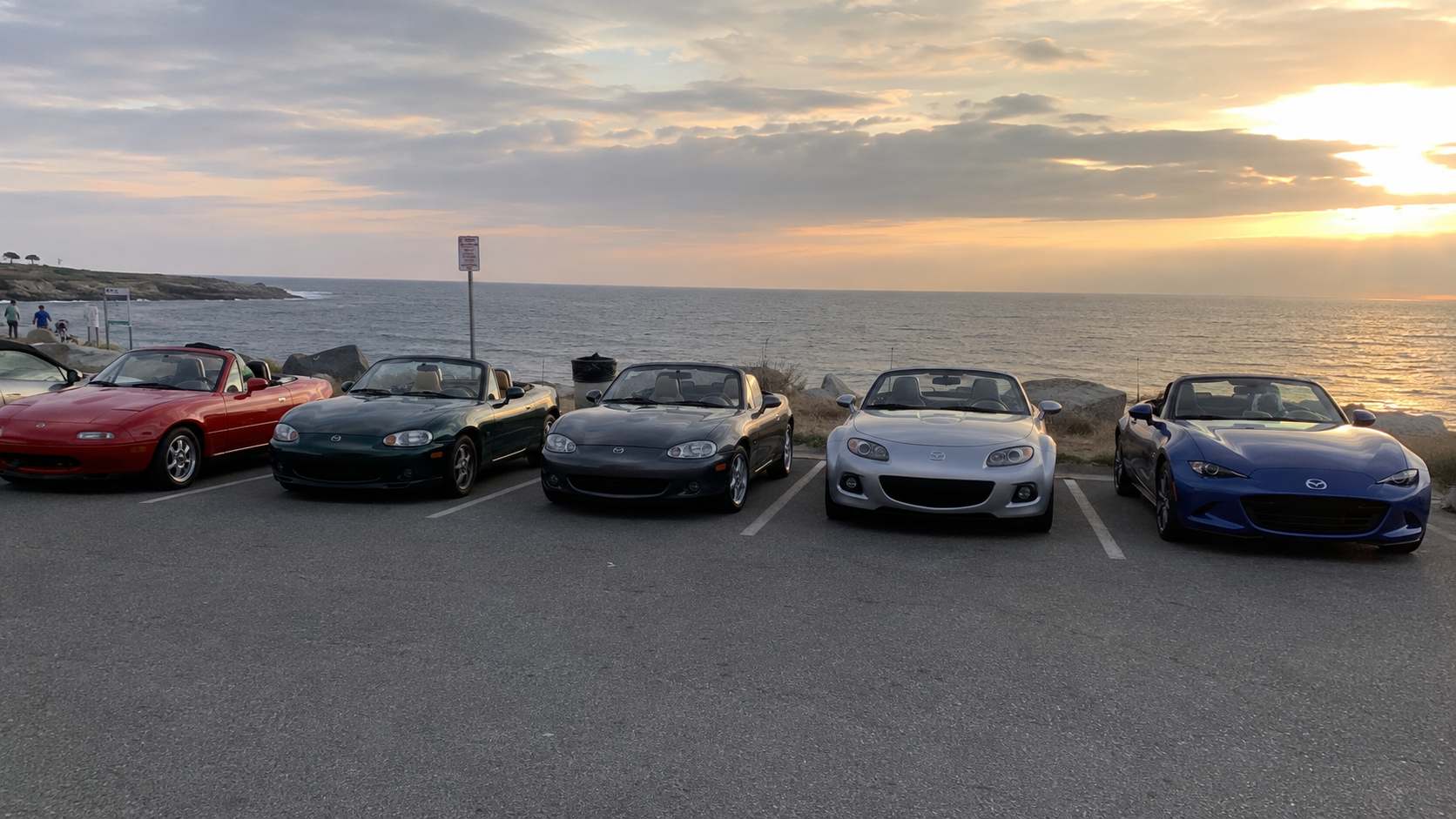 Cinq Mazda MX-5 de quatre générations alignées au bord de mer au coucher du soleil