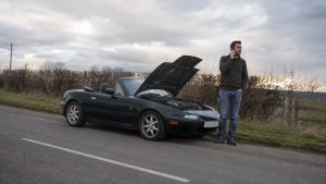 Mazda MX-5 NA verte en panne au bord d'une route de campagne, capot ouvert, conducteur au téléphone