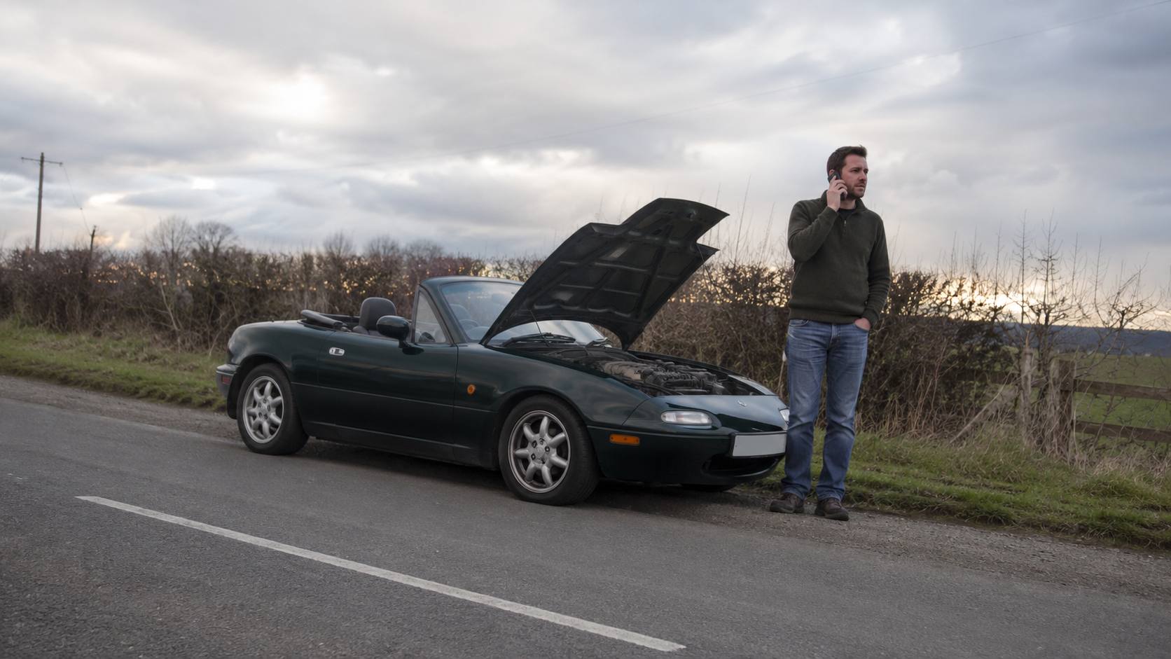 Mazda MX-5 NA verte en panne au bord d'une route de campagne, capot ouvert, conducteur au téléphone