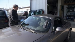 Mazda MX-5 cabriolet gris dans un atelier automobile en plein soleil, technicien avec gants examinant le pare-brise fissuré, plusieurs impacts visibles dans la partie supérieure du vitrage, ventouse professionnelle fixée sur le pare-brise, essuie-glaces et habitacle visibles, vitres automobiles stockées à proximité du garage, scène réaliste de diagnostic et réparation illustrant les pièges, le prix de remplacement et les conséquences au contrôle technique, juillet 2026