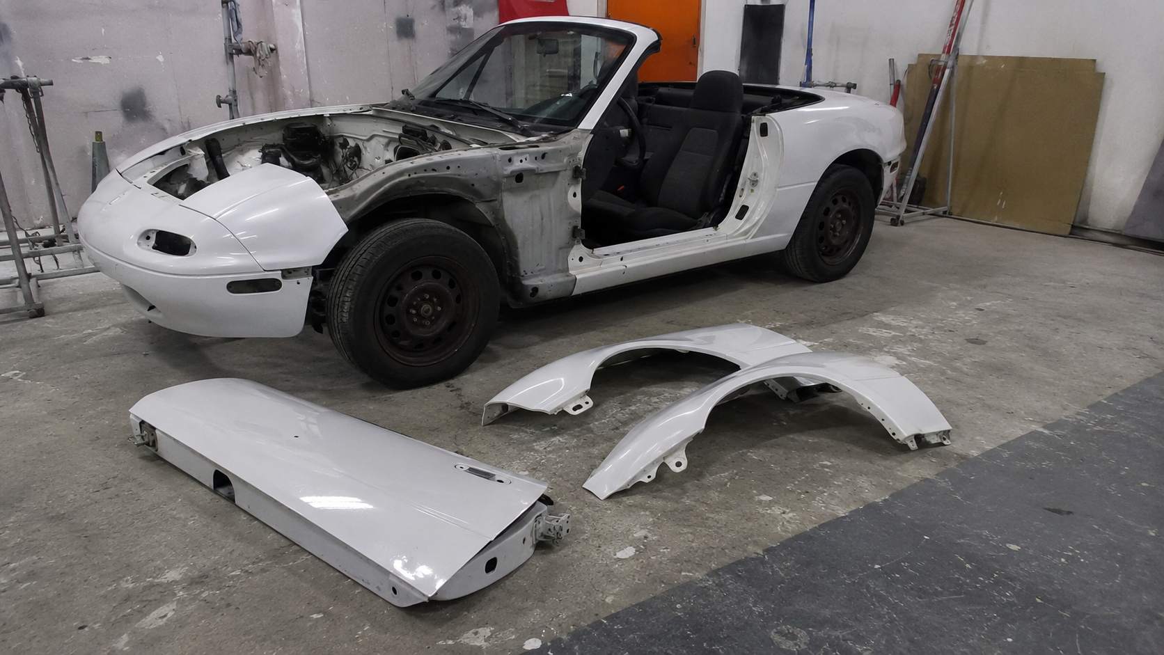 Mazda MX-5 NA blanche en restauration, ailes et portière déposées au sol d'une cabine de carrosserie