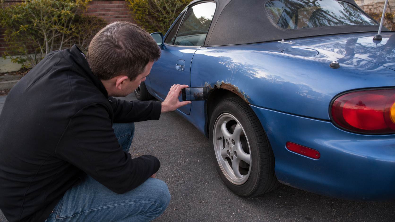 Homme photographiant au smartphone la rouille du passage de roue arrière d'une Mazda MX-5 NB bleue
