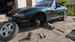 Mazda MX-5 NB verte sur cric devant un garage, roue avant déposée, moyeu et roulement de roue visibles, nouveau roulement posé au sol avec outils de mécanique, remplacement du roulement avant, lumière naturelle ensoleillée, 22 juillet 2026, format 16:9