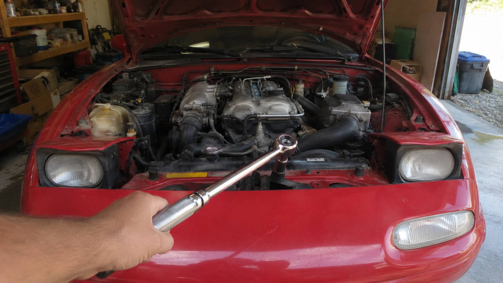 Main tenant une clé dynamométrique devant le moteur d'une Mazda MX-5 NA rouge aux phares escamotables ouverts