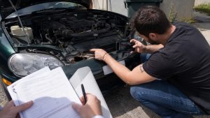 inspection d’une Mazda MX-5 verte au capot ouvert dans une cour ensoleillée, homme accroupi photographiant avec son smartphone une importante corrosion perforante sur la traverse avant près du phare, seconde personne tenant un dossier de documents et un stylo pour constituer les preuves d’un vice caché et demander le remboursement du véhicule, photographie documentaire réaliste Canon EOS R6, 27 juillet 2026, format 16:9