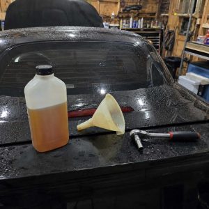 Photographie carrée de 1254 x 1254 px cadrée sur le capot de coffre noir brillant d'une Mazda MX-5, entièrement perlé de gouttes d'eau. Trois éléments posés dessus : bidon plastique translucide à bouchon noir rempli aux deux tiers d'une huile ambrée tirant sur l'orange, entonnoir souple jaune translucide couché sur le flanc, cliquet à douille chromé à manche bicolore noir et rouge équipé d'une douille longue. Au-dessus : lunette arrière en verre avec fils de dégivrage horizontaux, capote noire relevée, troisième feu stop rouge intégré au capot de coffre. Arrière-plan d'atelier à murs en panneaux de bois : étagère garnie d'aérosols et de bidons bleus, jaunes, blancs et rouges, servante à outils noire à tiroirs, faisceau de durites et de câbles noirs suspendus, conteneur à déchets noir, bacs plastique bleu et blanc, rayonnage métallique. Éclairage artificiel chaud, reflets spéculaires marqués sur la carrosserie humide.