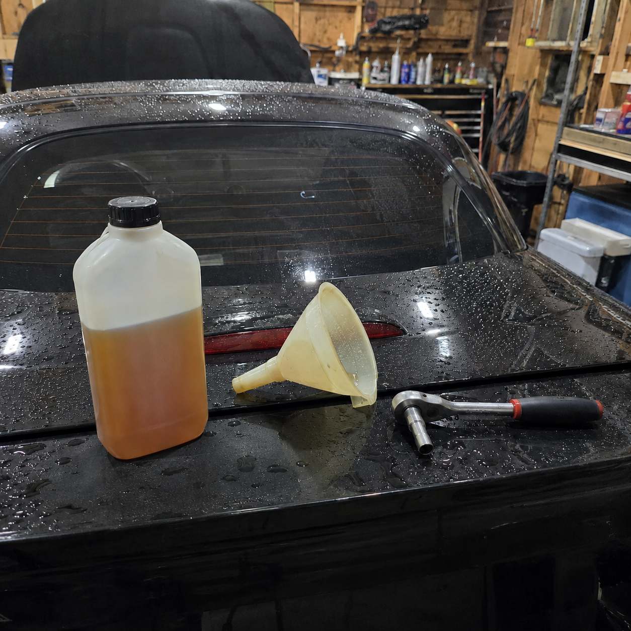 Bidon d'huile de pont ambrée, entonnoir jaune et cliquet posés sur le coffre noir d'une Mazda MX-5
