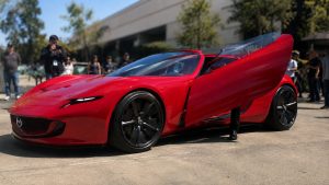 Mazda Iconic SP rouge vif photographiée en extérieur par temps ensoleillé, concept-car sportif très bas vu de trois quarts avant gauche, carrosserie aérodynamique, projecteurs LED fins, grandes jantes noires, porte droite spectaculaire ouverte vers le haut, habitacle partiellement visible, voiture nette au premier plan, spectateurs, arbres et bâtiment moderne fortement floutés par un bokeh naturel en arrière-plan, lumière directe et ombres réalistes, rendu photo documentaire grand-angle 16:9, 2026.