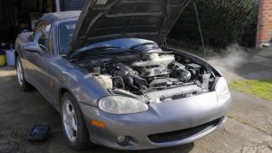 Mazda MX-5 NBFL grise de 2001 stationnée devant un garage en plein soleil, capot ouvert sur moteur essence, diagnostic de broutement à froid avec valise OBD et câbles au sol, contrôle admission allumage injection et capteurs moteur, légère vapeur visible à proximité, vue trois-quarts avant grand-angle 24–28 mm,
