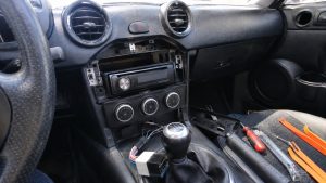 Autoradio Mazda MX-5 : bien choisir et l'installer sans erreur 2 Installation autoradio Mazda MX-5, console centrale démontée avec poste radio aftermarket, emplacement DIN ouvert, faisceau électrique multicolore et connecteur adaptateur visibles près du levier de vitesses, commandes de climatisation circulaires, outils de démontage orange et tournevis sur siège passager, intérieur noir éclairé par lumière naturelle ensoleillée, prise de vue grand-angle 24–28 mm Canon EOS R6, août 2026