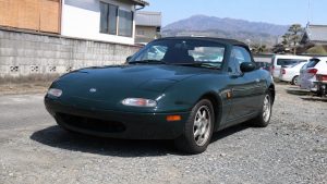 Eunos Roadster Mazda MX-5 NA japonaise vert foncé, cabriolet deux places avec capote noire, phares escamotables fermés, jantes alliage argent, vue trois quarts avant sur parking en gravier au Japon, maisons traditionnelles et montagnes en arrière-plan, lumière naturelle ensoleillée, photographie documentaire Canon EOS R6 grand-angle 24-28 mm, profondeur de champ modérée, textures réalistes, août 2026