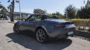 MX-5 propulsion ou traction : la réponse claire et sans détour 2 Mazda MX-5 ND cabriolet gris métallisé capote ouverte, vue arrière trois quarts côté conducteur, stationnée sur chaussée asphaltée en plein soleil, roues alliage multibranches visibles, roue arrière au premier plan, feux arrière circulaires rouges, double arceau derrière les sièges, échappement arrière droit, carrosserie réfléchissante, palmiers, haies et relief urbain en arrière-plan, photographie automobile documentaire grand-angle 24-28 mm, lumière naturelle dure, ombres nettes, illustrant la transmission propulsion de la MX-5, 2026