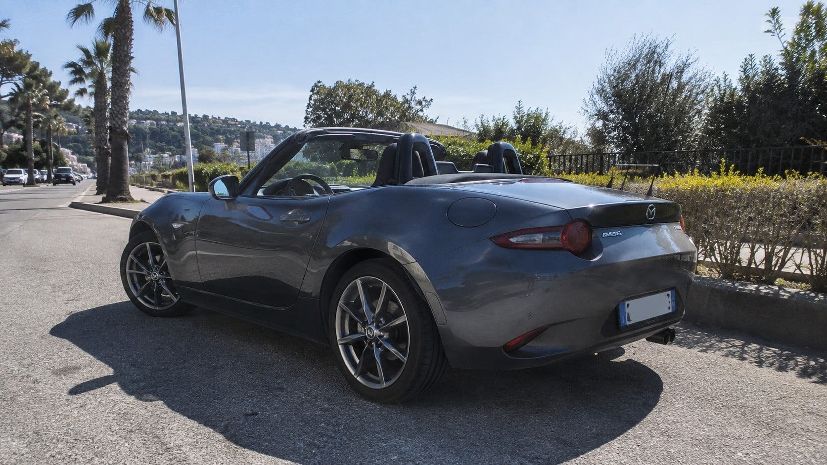 Mazda MX-5 ND cabriolet gris métallisé capote ouverte, vue arrière trois quarts côté conducteur, stationnée sur chaussée asphaltée en plein soleil, roues alliage multibranches visibles, roue arrière au premier plan, feux arrière circulaires rouges, double arceau derrière les sièges, échappement arrière droit, carrosserie réfléchissante, palmiers, haies et relief urbain en arrière-plan, photographie automobile documentaire grand-angle 24-28 mm, lumière naturelle dure, ombres nettes, illustrant la transmission propulsion de la MX-5, 2026