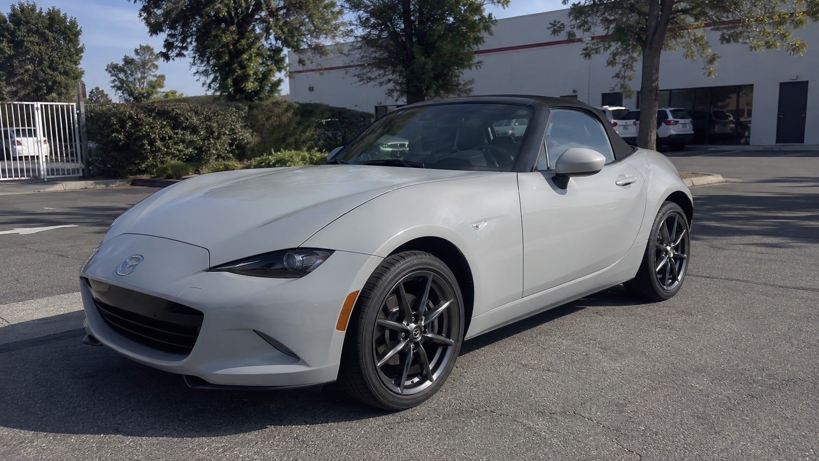 Mazda MX-5 neuve 2026 : prix, finitions et guide d'achat 1 Mazda MX-5 neuve 2026 argent métallisé, roadster deux places avec capote souple noire fermée, vue trois-quarts avant gauche, bouclier avant et grande calandre noire, optiques effilées, jantes alliage multibranches foncées, pneus visibles, carrosserie réfléchissant les arbres, véhicule stationné sur asphalté en extérieur, bâtiment commercial blanc et véhicules en arrière-plan, végétation et arbres, forte lumière naturelle ensoleillée, ombres réelles au sol, cadrage grand-angle documentaire 24–28 mm
