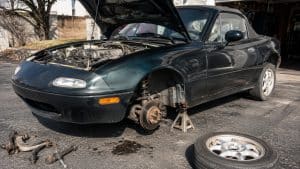 Mazda MX-5 NA cabriolet vert foncé immobilisée en extérieur au soleil, capot moteur ouvert, roue avant gauche démontée, disque et moyeu de frein visibles, véhicule posé sur chandelle, roue déposée au premier plan, bras et pièces de suspension démontés sur le sol, traces de liquide sous le train avant, scène de diagnostic mécanique illustrant les pannes connues et coûts de réparation Mazda MX-5, 2026
