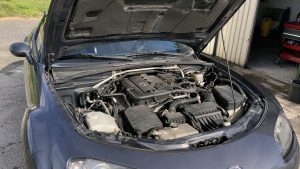 Fiabilité du moteur MX-5 NC 2.0 : ce qu'il faut savoir 1 Mazda_MX_5_NC_2_0_grise_capot_ouvert_vue_avant_compartiment_moteur_essence_bloc_moteur_collecteur_admission_durites_boite_a_air_reservoirs_liquides_jauge_huile_barre_renfort_elements_mecaniques_poussieres_usage_normal_stationnement_exterieur_devant_garage_lumiere_naturelle_ensoleillee_controle_fiabilite_moteur_aout_2026