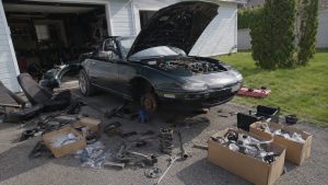 Restauration complète d’une Mazda MX-5 NB vert foncé immobilisée devant un garage sous un soleil marqué, capot moteur ouvert, roue avant droite démontée avec disque de frein apparent, véhicule posé sur chandelle, sièges retirés, ressorts de suspension, bras mécaniques, freins, pièces moteur, cartons de composants neufs, caisses et outils répartis sur l’allée, carrosserie avant usée et partiellement rayée, scène réaliste de rénovation automobile illustrant le budget poste par poste, photographie documentaire horizontale 16:9 au Canon EOS R6, objectif grand-angle 24 à 28 mm, lumière naturelle, profondeur de champ modérée, textures détaillées, août 2026