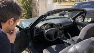Airbag Takata Mazda MX-5 : votre roadster est-il concerné ? 2 Mazda MX-5 cabriolet verte, portière conducteur ouverte, homme inspectant l’habitacle, volant avec module airbag conducteur central, combiné d’instrumentation à cadrans blancs, planche de bord noire, boîte manuelle, frein à main, sièges tissu, capote ouverte, stationnement résidentiel en plein soleil